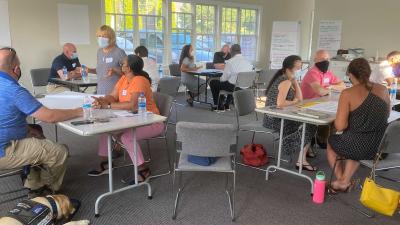 Diverse group of community members working animatedly in small groups. Police dog naps in corner.